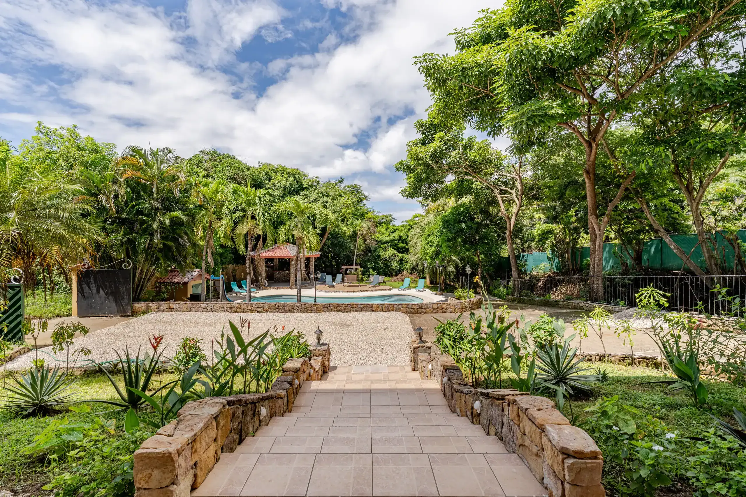 Pathway leading from the main gate to the house entrance, lined with greenery and open views of the pool area.